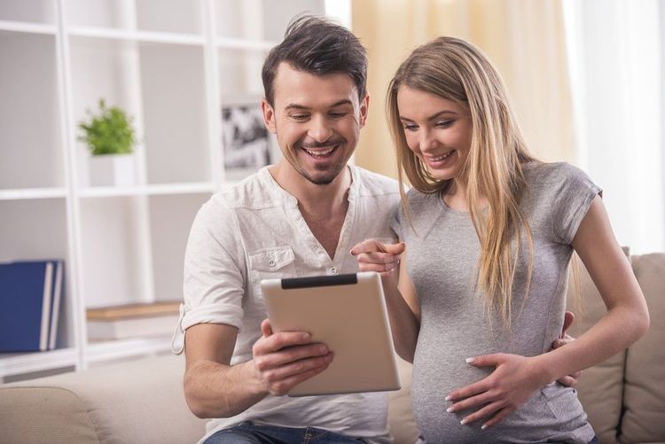 Smiling pregnant woman sitting on a blanket in a park, holding a tablet for a video call.