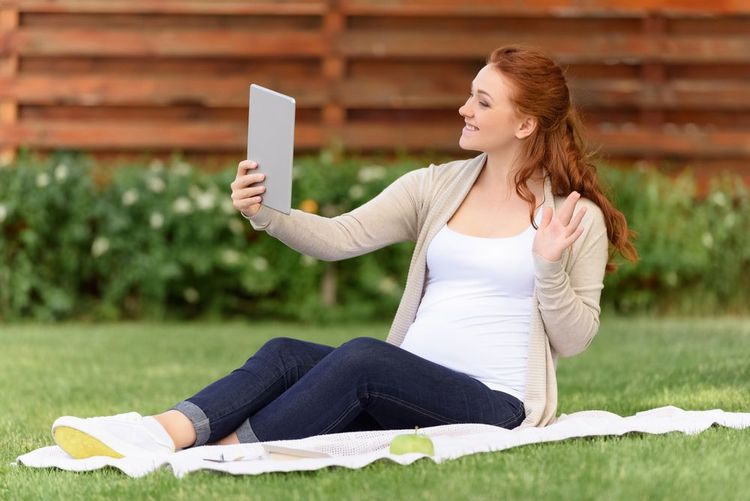 Smiling pregnant woman sitting on a blanket in a park, holding a tablet for a video call.