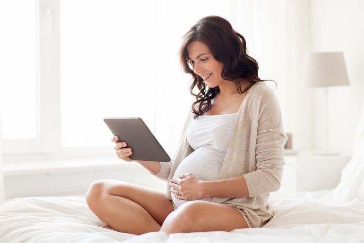 Smiling pregnant woman sitting on a blanket in a park, holding a tablet for a video call.