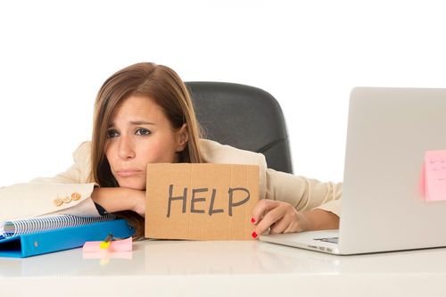 Woman sitting at a desk looking stressed, holding a cardboard sign with the word "HELP" in front of a laptop.