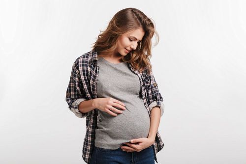 Pregnant woman standing and looking down at her belly, wearing a plaid shirt and grey top.