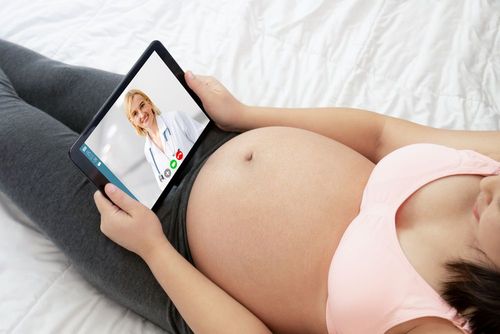 Pregnant woman lying on bed, holding a tablet and having a video call with a doctor.